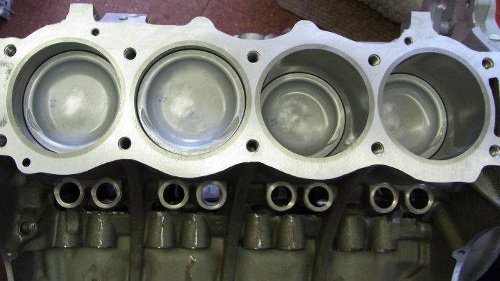 Pistonheads Upgrade - The image features an engine block with individual cylinders. The cylinders are lined up in a row, indicating a V-shaped layout where two rows of cylinders meet at the center. Each cylinder appears to have a bolt pattern to attach components such as pistons and a block cover. There are six main cylinders in the foreground, while the bodies of the cylinders are partially visible. The metal texture of the engine suggests a sturdy construction, typical for vehicle engines. The perspective is a top-down view, though the engine is slightly tilted, giving a sense of depth.