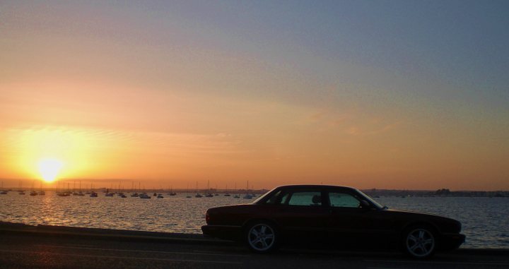 Jaguar Pistonheads Xjr - The image captures a serene sunset scene on a waterfront. A vintage car is parked on a concrete ledge overlooking a marina filled with boats. The setting sun casts a soft orange glow on the car and the still water, reflecting the last rays of the day. The image invokes a sense of tranquility and timelessness.