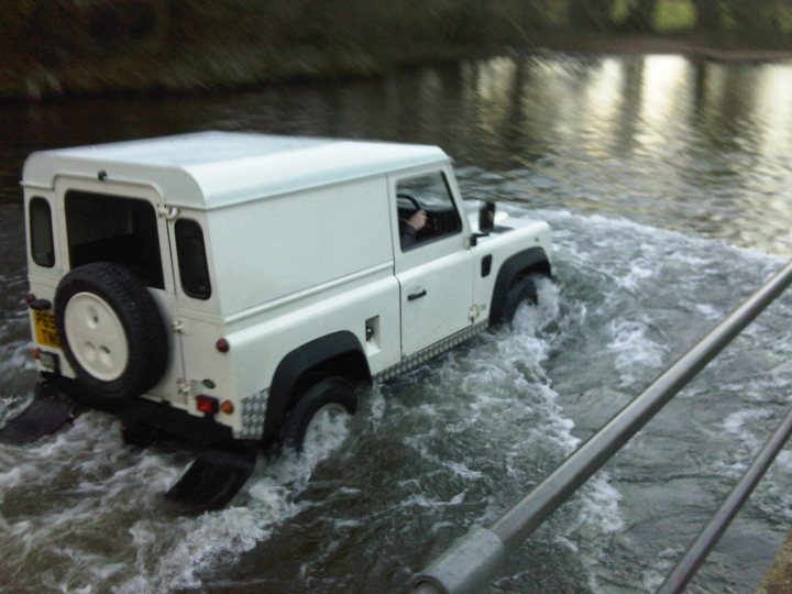 Pistonheads - The image depicts a white vehicle, appearing to be a truck, driving through a flooded area. The vehicle is partially submerged and is navigating through what might be a former road or bridge. The floodwaters cover the ground surrounding the truck, indicating a significant volume of water. A railing is visible along the right edge of the image, suggesting this scene is taking place near a man-made structure. The atmosphere of the image conveys a sense of urgency or emergency, possibly arising from extreme weather conditions or natural disasters.