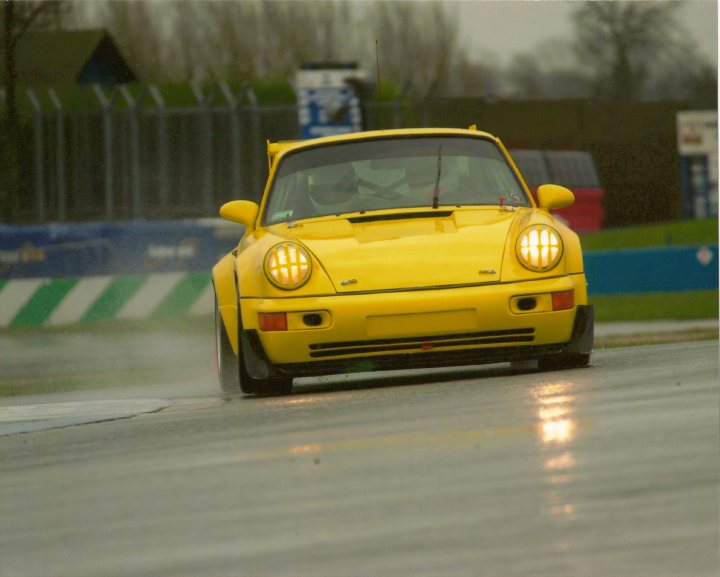 Pistonheads - The image captures a vibrant scene from a rally race. At the heart of the action is a **yellow racing car** with a **black hood**, skillfully navigating a wet road. The car is leaning to one side, suggesting a sharp turn, and the driver's side window is open, perhaps for better aerodynamics or to let in some breeze. The car is moving parallel to a **blue guardrail**, indicating the presence of a track guide or a safety barrier. The background of the image reveals more of the race track, with a **blue building** visible in the distance, adding a splash of color to the scene.