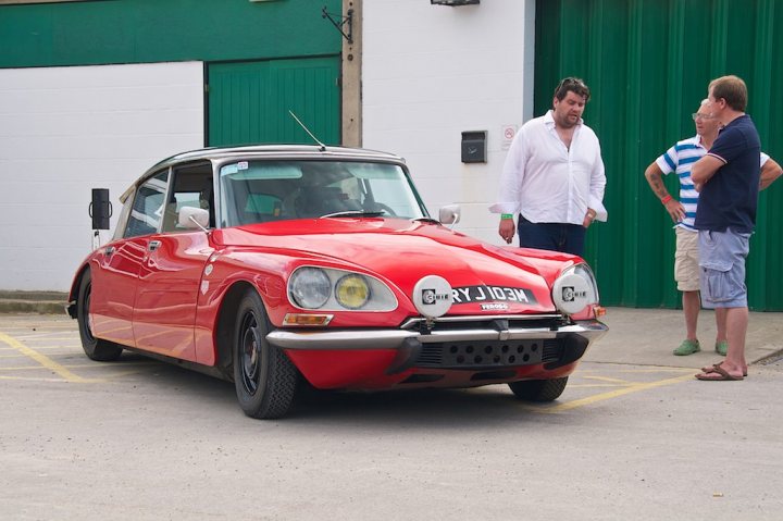 Tyre advice from Longstone Tyres - Page 1 - Classic Cars and Yesterday's Heroes - PistonHeads - In the image, there is a group of people gathered around a vintage red car parked on the side of a street by a building with a green door. Two men are standing on the sidewalk, engaged in conversation, while a woman and another man are positioned slightly behind the car. The car appears to be a sports model, judging by its sleek design and angular features. It's a bright, sunny day, suggesting it's summer or early spring.