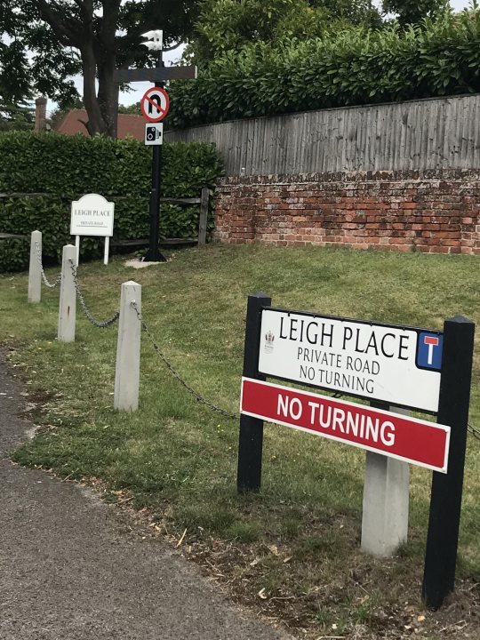 What’s the legal take of no turning signs on private roads  - Page 1 - Speed, Plod & the Law - PistonHeads - The image is a photograph of a road sign with the text "Leigh Place" visible. It appears to be taken during daylight under overcast or cloudy skies. In the background, there is a hedge and a stone wall, suggesting a suburban or rural location. There are also other signs in the vicinity indicating parking restrictions and possibly speed limits, which are common elements of roadside signage. The style of the image is candid and appears to be taken by an amateur photographer rather than a professional one.