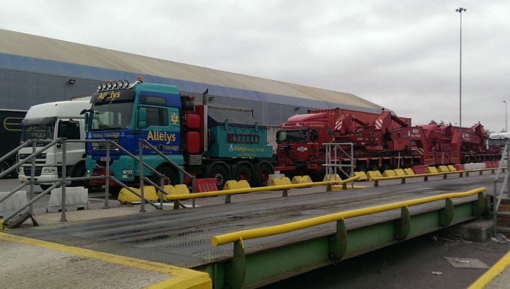 The Heavy Haulage Appreciation Thread - Page 12 - Commercial Break - PistonHeads - The image shows a loading dock with two trucks parked. On the left, there is a white truck with a large space for a trailer, and on the right, there is a red truck pulling a large trailer. Behind these vehicles, there is a large building with a distinctive blue-striped awning that dominates the background. The dock itself seems to be in an industrial area, equipped with yellow loading equipment and safety barriers. The weather appears to be overcast.