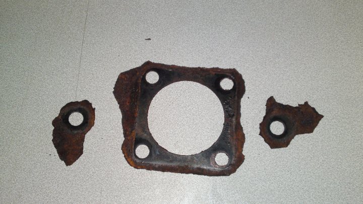 Project Peterborough - Page 3 - Alfa Romeo, Fiat & Lancia - PistonHeads - The image displays rusted metal components on a surface. These parts appear to be the remains of a mechanical assembly, possibly a flap from a vehicle's dump valve or a piece of the engine bumper. The rust has significantly deteriorated the surface of the metal, indicating exposure to moisture and oxygen over time. Two intact holes are clearly visible, suggesting that these parts are not whole but fragments from a larger assembly. The metal exhibits various hues of rust, suggesting a process of corrosion at different stages. The surrounding surface appears to be a grey, non-porous material, which contrasts with the reddish-brown color of the rust.