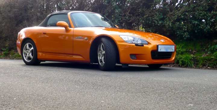 Rice Pistonheads Burner - The image shows an orange car parked on a paved street. The car is sleek and modern, suggesting it might be a sports car or a convertible. The vehicle is stationary with wheels turned towards the curb, indicating it is parked legally. The road on which the car is parked stretches into the distance with greenery visible on the margins.