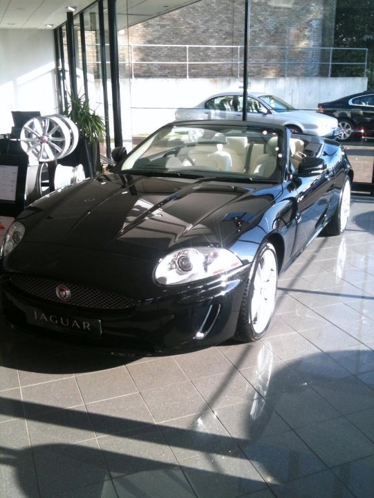 2011 XK 5.0 Convertable - Page 1 - South West - PistonHeads - This is a photograph of a black luxury sports car on display inside a spacious dealership showroom. The car's sleek design and shiny finish reflect its striking presence. It's positioned at an angle to the camera, giving a clear view of its front and side. The floor appears to be a shiny, polished tile, and gently scattered light makes the vehicle's features stand out against the room's ambient lighting.