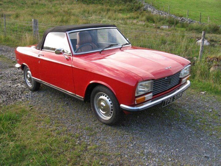 Lets see a picture of your classic(s) - Page 121 - Classic Cars and Yesterday's Heroes - PistonHeads - The image shows a vintage red car parked on a gravel road. It has a convertible top and is positioned in what appears to be a rural or outdoor setting, with lush green grass around it. There's a fence bordering the road on one side, and the car is facing towards the viewer. The car's design suggests it's an older model, and it's daylight, likely during the spring or summer season.