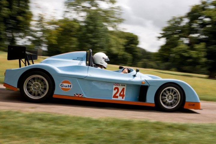 Motors Pistonheads Manor - The image portrays a thrilling scene on a race track. A blue sports car, emblazoned with the number 24 and the sponsor "Gulf," is in motion. The driver, dressed in a white helmet and a black suit, is intensely focused on the race ahead. The car is seen from a rear three-quarter perspective, giving a sense of speed and direction. The background reveals a grassy area, with trees lining the track, adding to the excitement and adrenaline of the race.