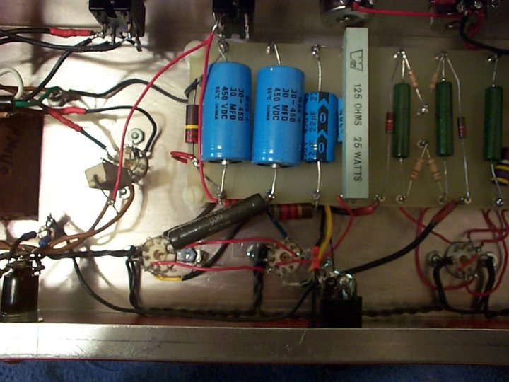 The image displays an intricate circuit board filled with an assortment of electronic components and wires. The board houses three capacitors, each labeled with the model number '25 WATS' and '12V.' The components are arranged in a structured manner, with resistors and a transformer interspersed among the capacitors. Several wires of varying lengths crisscross across the board, connecting the components and forming a complex network. The background is neutral, allowing the vibrant colors and precise arrangement of the components to stand out.