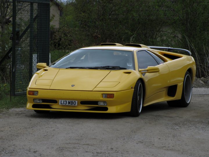 Pistonheads - The image depicts a yellow Lamborghini sports car, characterized by its powerful design and distinct branding. The car is parked on a gravel surface, suggesting an outdoor or off-road setting. It's positioned facing the camera, giving a clear view of the front and side of the vehicle. Behind the car, there's a green chain link fence and a partially visible white building, implying a residential or commercial area. The setting appears quiet, and the car seems to be the primary subject of the photograph.