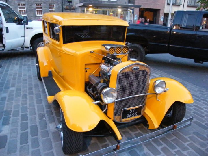 Edinburgh Cruisemeet Pistonheads - The image showcases a multi-colored classic car, painted a vibrant yellow with rust colored detailing accented by gold stripes. The car's robust engine takes center stage due to the distinctive hood that proudly displays it. The vehicle is parked on a cobblestone street, suggesting an older part of a city or town, and newer vehicles are visible in the background, providing a contrast between the old and the new. The car is a vintage model, filled with character and grit.