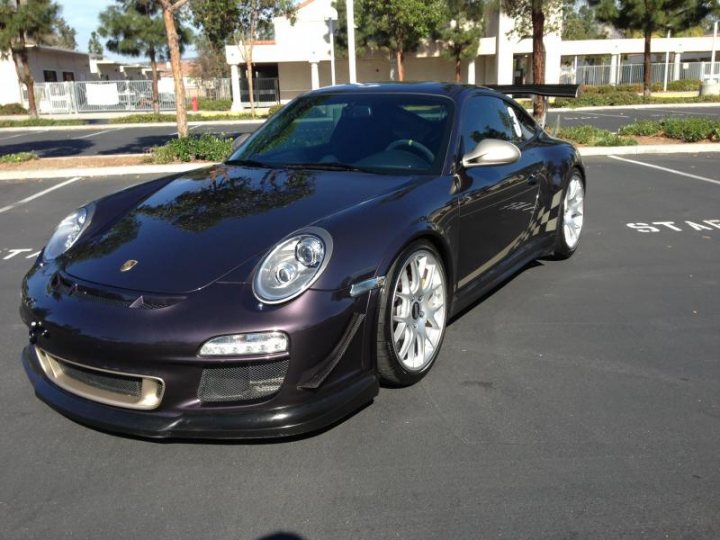 Porsche "Colour to Sample" Examples - Page 11 - Porsche General - PistonHeads - The image showcases a sleek Porsche Cayman sports car parked in a black parking lot. The car is impeccably shiny, reflecting the brick pavement and trees around it. It features a prominent front grille, a distinctive rear spoiler, and black rims encircling the tires. The side mirrors and handle are finished in shiny chrome. The vehicle is parked near a building, possibly in front of its entrance, suggesting the car might be the owner's mode of transportation.