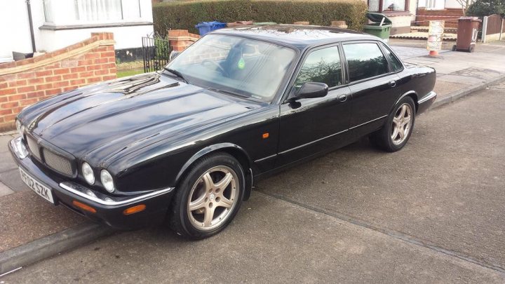 Pistonheads - The image shows a three-series BMW parked on a street next to a driveway. The car is predominantly black, with the exception of the silver wheels. It is a four-door model with the characteristic BMW design elements. On the driveway, there is a wooden fence and a blue trash bin. The photo appears to be taken during the day under clear skies.