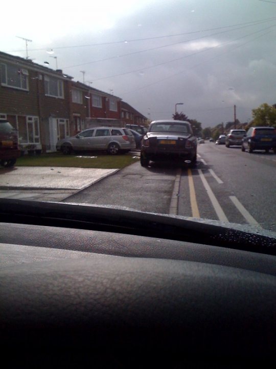 Tbops Phantom Pistonheads - The image captures a bustling urban scene viewed from a moving car. It's a cloudy day, and the car dashboard reflection shows on the windshield. The road is lined with parked cars, and there are six cars visible, including a silver van and a brown car. The buildings in the background appear to be residential white apartment houses. The street is paved and devoid of any visible pedestrians or bikers, creating an impression of calm under the overcast sky.