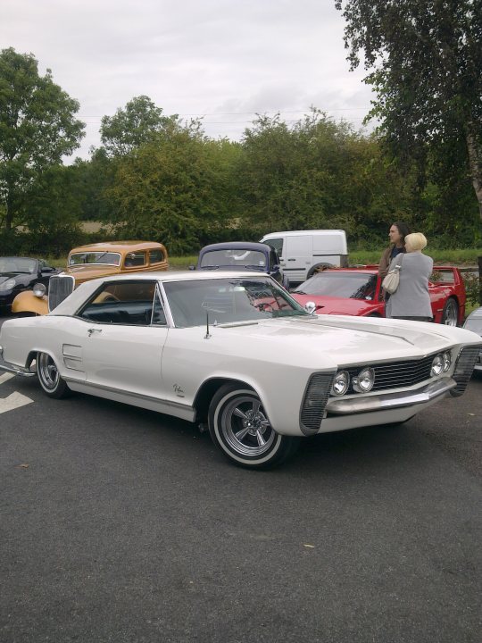 Pistonheads - The image features a pristine white classic muscle car, likely a 1969 Pontiac GTO, parked on a road next to several other vintage cars. The muscle car stands out due to its distinctive design and robust size, compared to the smaller vehicles surrounding it. The background is populated with trees and a clear sky, providing a serene setting for this gathering of classic automobiles. A few people can be seen near the edge of the frame, possibly admiring the vintage cars or preparing for a drive. The overall scene conveys a sense of nostalgia and appreciation for the aesthetics and history of these classic vehicles.