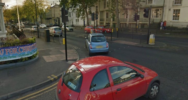 A red car driving down a street next to tall buildings - Pistonheads