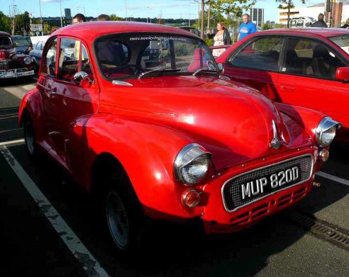 Pistonheads - The image showcases a parking lot scene featuring a striking red classic car positioned in the center of the frame. This car, with a license plate reading "MP 820", is the focal point of the image, drawing attention due to its vibrant color and the contrast between the surrounding vehicles and the asphalt background. Other vehicles, including a blue car visible behind the red classic car, are present in the background but are significantly less prominent than the main subject. The setting appears to be a wide paved parking area, with a few individuals scattered throughout, possibly pedestrians on the street or other visitors to the location.