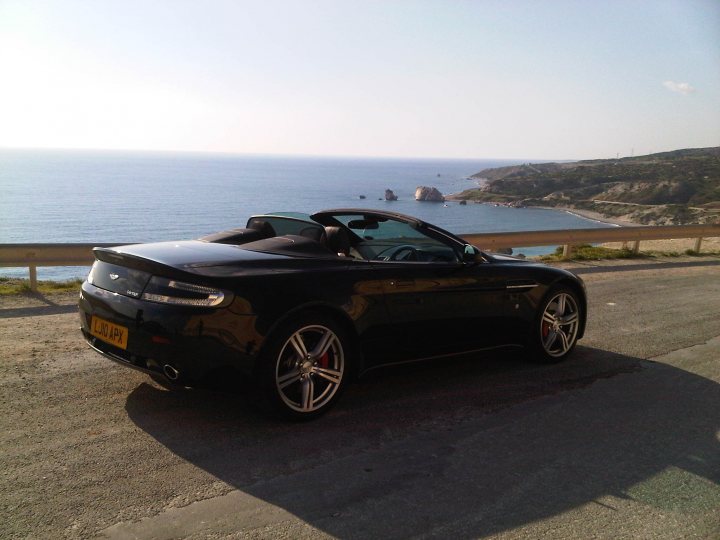 Hello from Cyprus! - Page 1 - Aston Martin - PistonHeads - In the image, a black sports car is parked along a coastal highway, overlooking the ocean and rocks. The car is situated on the right side of the road, which is marked by a guardrail. The backdrop of a clear sky, sea, and distant mountains adds to the serene and picturesque effect. A lone figure can be seen on the cliff above, adding a sense of scale and liveliness to the tranquil scene. The overall mood of the image is one of peace and freedom, as suggested by the open road and the vast expanse of the natural environment.