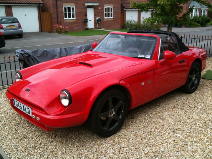 Honest Opinions Pistonheads - The image shows a vibrant red sports car parked beside a gravel driveway. The vehicle's sleek design and shiny finish reflect the sun, indicating fair weather. The setting appears to be residential, with a brick house visible in the background and a neatly maintained hedge lining the driveway. The gravel driveway descends slightly towards a gate in the background. There is also a glimpse of other cars in the vicinity, suggesting this might be a shared parking space. The car's vintage style, with its round headlights and distinct wheel design, adds a classic touch to the overall scene.