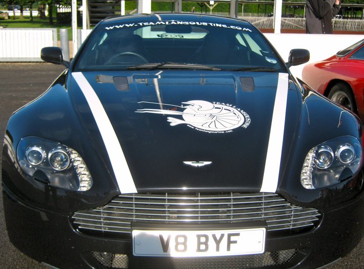 Pistonheads Stickered - The image showcases a sleek black sports car parked against a blurred backdrop, emphasizing the car's design and features. The front grille is prominently visible with the Aston Martin logo in the center, indicative of the car's luxury brand. A distinctive racing team-inspired stripe adorns the hood, adding to the car's sporty appeal. The license plate reads "V8 BYF," aligning with the car's sporty features. The rear view with other cars partially visible indicates it may be in a parking lot or a showroom.