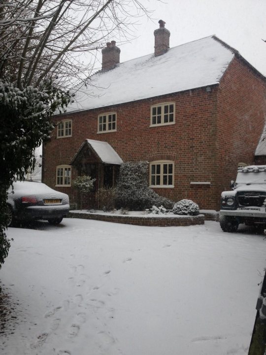 A red fire hydrant sitting in the middle of a street - Pistonheads - The image portrays a serene winter scene. A two-story house with a dark roof blanketed in snow serves as the main focus. The house features a smaller extension on the left side, also coated in a layer of snow. A driveway, flanked by stone walls, is visible in the snow-covered foreground, leading the eye towards the house. The ground is covered in snow, with footprints leaving a distinctive mark in the pristine white blanket. To the right, a few parked vehicles are covered in snow, adding to the wintry ambiance. The overall image presents a calm and quiet afternoon under snowy conditions.