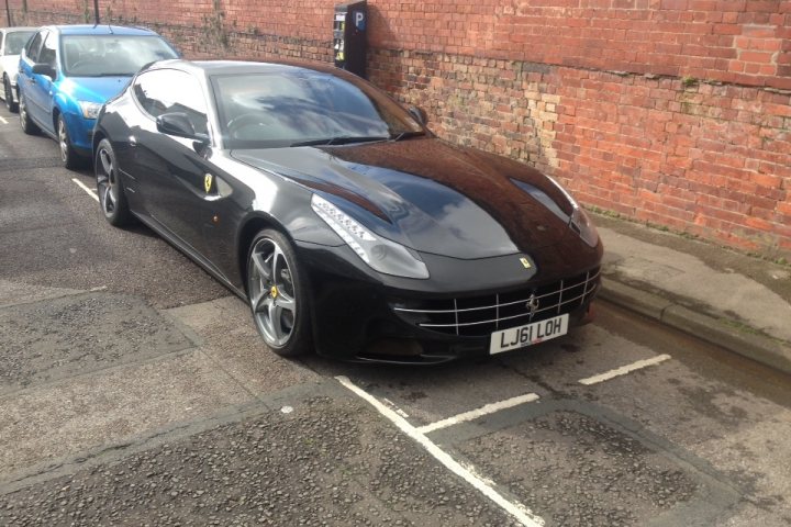 Tragic Ferrari FF.... - Page 1 - Ferrari V12 - PistonHeads - This image features a black Ferrari parked on the side of a road between two other vehicles. The Ferrari, a luxury sports car, is the central focus of the image. The license plate of the Ferrari reads "LJS LH," providing some detail about the vehicle. The Ferrari is parked in front of a brick wall, which suggests an urban environment. The day is partly cloudy, contributing to the overall atmosphere of the scene.