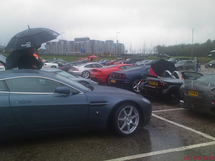 Pistonheads - The image showcases a large parking lot filled with a variety of beautifully designed cars. The cars are parked haphazardly, giving the impression of a busy event. Among the gray and silver cars, a few red and black ones stand out. In the foreground, a person is seen holding an umbrella, possibly indicating recent rain or the coming of precipitation. The sky overhead is overcast, casting a soft, diffuse light throughout the scene.