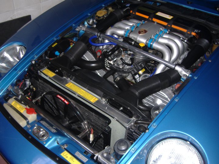 Porsche 928 40th anniversary in 2017 - Page 1 - Front Engined Porsches - PistonHeads - The image depicts the engine bay of a blue luxury car. Various components of the engine are visible, including tubes that run diagonally towards the front of the car. The engine bears the Porsche logo, indicating it is a Porsche model. The hood is open, revealing the engine's complexity and the presence of multiple filters. The presence of caution tape suggests a focus on safety while observing or working on the vehicle's engine.