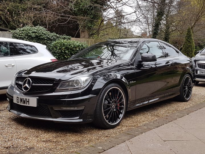 What car for a Bank Robbery ?? - Page 4 - The Lounge - PistonHeads - The image shows a black Mercedes sports coupe parked on a gravel driveway. The car has a sleek design with a large front grille featuring the Mercedes emblem. The vehicle is equipped with five-spoke black wheels and distinctive side mirrors. The surrounding environment includes trees, a fence, and the rear end of another vehicle parked nearby. The sky is overcast, suggesting a cloudy day.