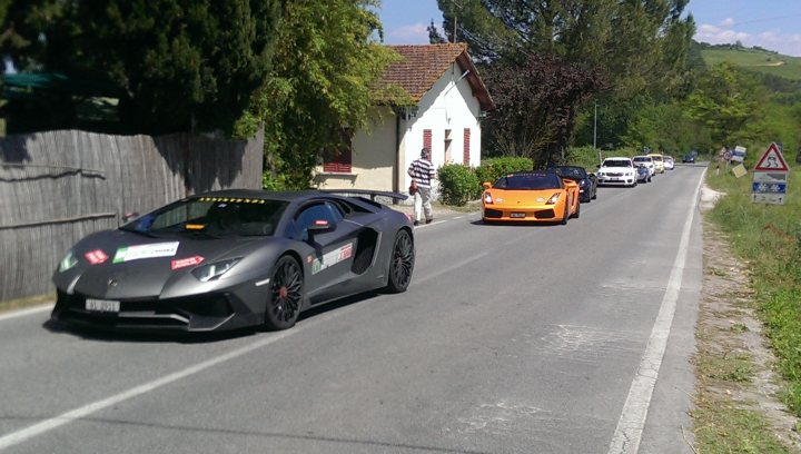 Supercars spotted, some rarities (vol 6) - Page 497 - General Gassing - PistonHeads - The image captures a thrilling scene on a winding mountainous road. Two incredibly fast sports cars, one shaded in black and the other in orange, are racing neck and neck. The driver of the orange car has his hair standing on end from the speed and excitement. A scenic backdrop of mountains and a clear sky adds to the drama and adrenaline of the scene. This image conveys the joy of speed and the beauty of nature.