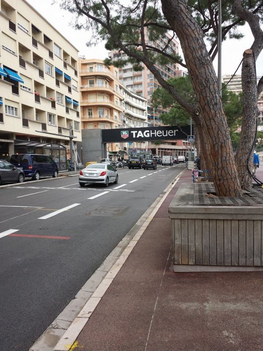 A city street filled with lots of traffic - Pistonheads - The image shows a city street scene with tall buildings lining one side. In the center of the image, a silver sports car is driving down the road, passing by a sidewalk with a bench. On the sidewalk, there is a tree with a member of the public standing next to it, facing the car. The street is well-maintained with clear lane markings. Visible storefronts indicate commercial activity.