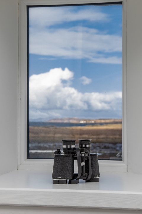 My wee Isle of Skye renovation - Page 7 - Homes, Gardens and DIY - PistonHeads UK - The image displays a view from a window with a white frame. Outside the window, there is a landscape featuring a coastline or perhaps an island in the distance, under a clear sky with some clouds. On a window sill inside, there are several binoculars standing next to each other. The room appears to be well-lit and has a plain wall in the background. There's also a small window on that wall.