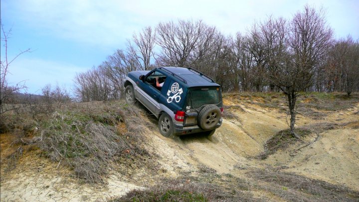 Couple of pics from today... - Page 1 - Off Road - PistonHeads - The image captures an exhilarating scene of off-road driving on a sandy hill. A person is seen driving a four-wheel drive vehicle, which is a mix of a dark blue car styled like a truck, equipped with off-road tires. The vehicle is tilted to one side, tilted considerably, displaying its capability to handle steep slopes. The terrain is rugged, with dry dirt and sandy patches, indicative of a challenging off-road track. The car is spewing dust behind it, demonstrating the vigorous performance of the vehicle in such conditions. The driver appears focused, gripping the steering wheel in anticipation of the next challenging move.