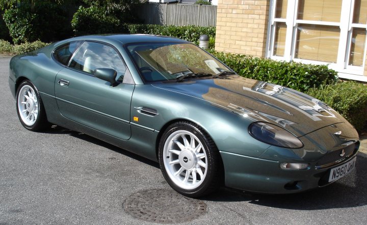 Your DB7 pictures. - Page 1 - Aston Martin - PistonHeads - The image showcases a car, specifically the frontal view of it parked in front of a building. The car is a darker shade of green, featuring a rounded body with a sleek, shiny finish. The bridge of a nearby water body can be seen in the background, adding to the aesthetics of the scene. A sewer grate is visible before the front wheel of the car, indicating the car is parked on a road that runs alongside the water. The overall impression is one of a serene and picturesque location.