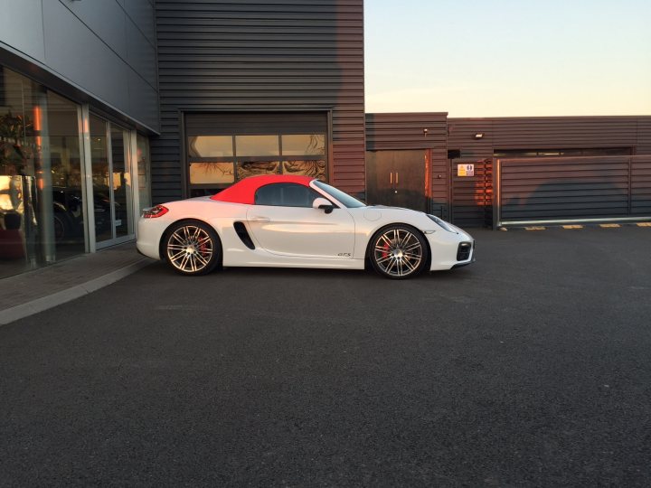 Cayman GTS - Page 110 - Boxster/Cayman - PistonHeads - The image showcases a modern, white sports car with a red rear wing parked in front of a building. The car appears to be a Porsche, judging by its distinctive design elements and the emblem visible on the side. The Porsche is parked on an asphalt surface, precisely in front of a set of large glass doors that lead into the building. The building itself has a contemporary style, with black metal cladding, large windows, and a dark gray roof. The backdrop is graced by a clear blue sky, suggesting a bright and sunny day. There's no mention of a second car, so it could be assumed that the scene includes only one car and building.