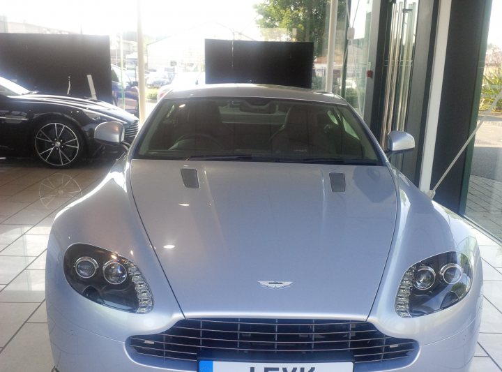 V8VS Lightning Silver with Pics - Page 1 - Aston Martin - PistonHeads - The image shows a shiny silver Aston Martin sports car parked inside a car showroom. The car is positioned facing to the right, with the hood slightly raised to reveal part of the sleek engine. The interior lighting reflects off the car's shiny surface, enhancing its metallic sheen. The showroom's floor and walls can be seen in the background, highlighting the exclusivity of the car showcased.