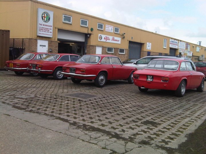 Pistonheads Bertones Beautiful - The image showcases a commercial building with a prominent entrance and sign, partially obscured. In front of the building, there is a collection of vintage cars, primarily red, parked on a cobblestone surface. These cars appear to be classic models, possibly sports or luxury cars from the mid-20th century. The presence of a metallic gate suggests a secure parking area or a gated property. The overall scene suggests a place associated with vehicle maintenance or historical displays of automobiles.