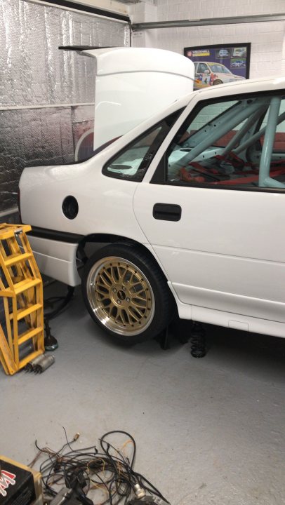 Vauxhall Cavalier GSI RestoMod Track Car - Page 16 - Readers' Cars - PistonHeads UK - The image is a photograph of the interior of a garage, showcasing a white car in the foreground that appears to be a rally or race car given its modified suspension and body kit. The vehicle has a prominent rear wing, a feature common in high-performance motorsport cars. To the right of the car, there is a yellow step ladder leaning against it, possibly for maintenance or repair work.

The garage itself is white with concrete floors, and there are some tools scattered around. In the background, you can see additional vehicles, suggesting that this space is used for multiple purposes beyond just racing cars. There's also a person visible on the left side of the image, though their role or activity in the scene is not clear from this perspective.

The photograph has a watermark or logo overlaying it, which seems to be related to a car restoration or customization service. This suggests that the image might have been taken during a visit to such a business for vehicle work or consultation.