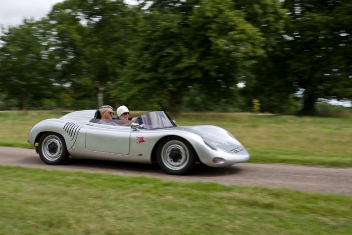 Manor Motors Pistonheads - The image showcases a vintage, open-top car with two occupants, both donning white knit hats and sunglasses, driving down a rural road flanked by trees. The car is gray and appears to be a classic model, distinct by its geometric patterns. The setting suggests a sunny day, with the lush greenery adding a serene backdrop to the car's journey. The couple seems to be enjoying their ride, immersed in the experience of cruising through the countryside in a stylish classic car.