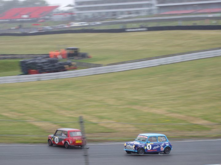 BRSCC Meeting Brands Hatch 11-06-2011 - Page 1 - UK Club Motorsport - PistonHeads - The image captures an exciting moment on a race track in England. Two small cars, one red and the other blue, are locked in a thrilling race around the curve of the track. The red car is positioned slightly behind the blue car, indicating a neck-and-neck competition. The backdrop of the track is lush with green, providing a stark contrast to the action on the track. The race track appears empty, with no other cars visible, offering a clear view of the ongoing competition between the two cars.