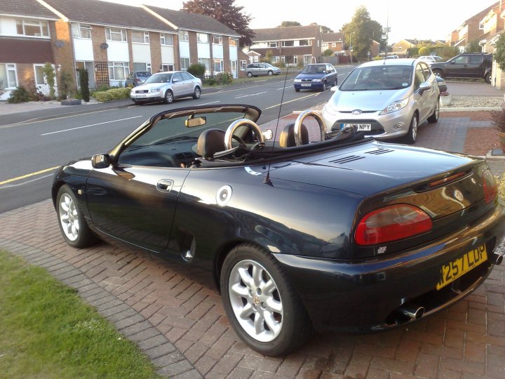 Hurt Pistonheads - This image shows a black convertible car parked on the side of a street. The car is styled with a design that suggests it's an older model. It has its top down, revealing two seats visible in the image. The vehicle is sleek and glossy, reflecting its surroundings. The street has parked cars on both sides, creating a tunnel effect. There are houses visible in the background, indicating a residential neighborhood. The sky appears to be cloudy, suggesting an overcast day.