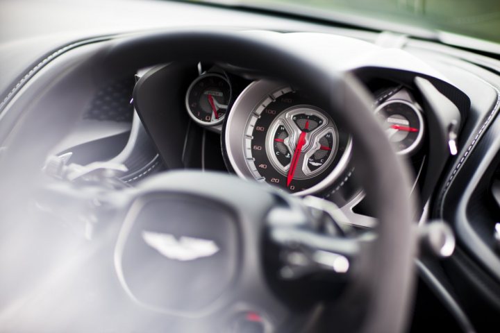 New DB10 Pictures Released - Page 1 - Aston Martin - PistonHeads - The image offers a driver's perspective from behind the wheel of a car. The interior is out of focus, allowing the viewer to focus on the dashboard and the circular dashboard gauge with red hands and black numbers. Tilted slightly to the left, the steering wheel, similar in design to those found in luxury or racing cars, is visible. The gear shift, identified by its setting between the steering wheel and the dashboard, is partially visible. The overall styling suggest a high-end or performance vehicle.