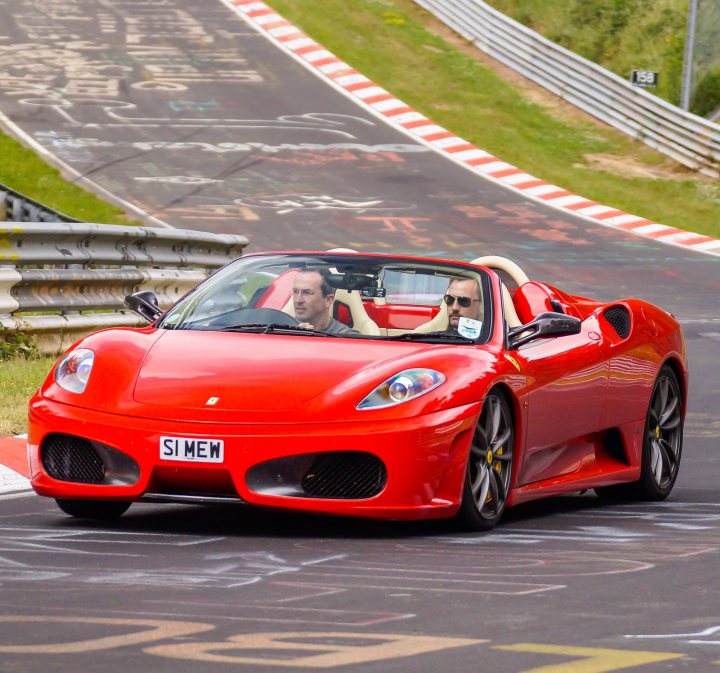 Ferrari F430 SPIDER F1 16M STYLING - Page 1 - Ferrari V8 - PistonHeads - The image depicts a lively race track scene, featuring a striking red sports car in motion. The car, which is a Ferrari, is painted a vibrant red with distinguishing white and black stripes on the rear wing. Two individuals are present in the car, both wearing white helmets and sunglasses, indicating they are likely the driver and passenger. The track itself is marked with a multitude of white lines, creating various specified lanes for vehicles. The setting suggests a competitive environment, with multiple spots for spectators or other racers.