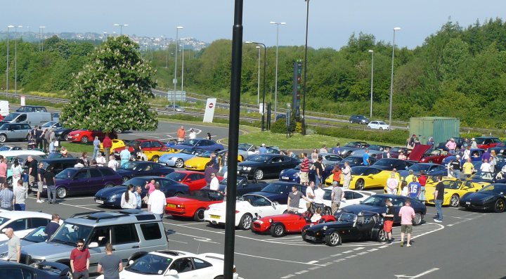 Gordano this morning en route to cwc - Page 1 - South West - PistonHeads - The image captures a bustling scene in a parking lot filled with various cars. The lot is packed with a diverse range of vehicles, including small cars, trucks, and even a bus, hinting at a large gathering or event. People are scattered throughout the lot, some near their vehicles while others seem to be mingling, suggesting a social atmosphere. The lot is surrounded by a lush green courtyard, adding a touch of nature to the scene. Above, the sky is mostly clear, indicating good weather for the day.