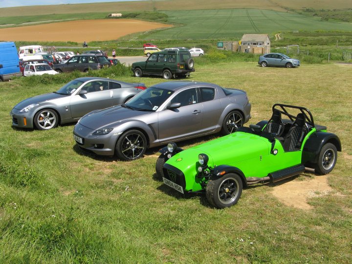 Photography Pistonheads South Coast - The image captures a vibrant scene on a grassy field next to a dirt road. Three cars are parked there, each uniquely styled. One is a sleek, silver sedan parked towards the left side of the image. The right side of the image features a bright, lime-green buggy with two seats, and next to it, a black-trimmed buggy with four seats, which could be a convertible. The vehicles are stationary, and the people near them appear to be engaged in conversation. In the background, a warm-colored landscape stretches out, suggesting a rural setting.