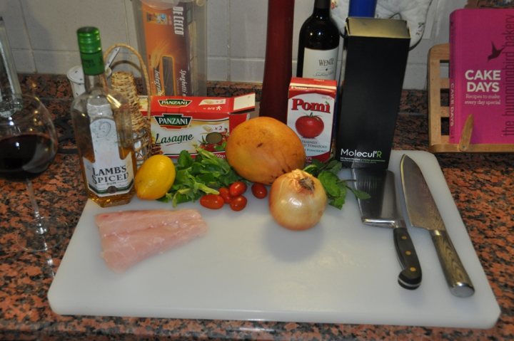 PH Cooking Competition #19 - A Twist on Tradition - Page 2 - Food, Drink & Restaurants - PistonHeads - This image shows a cutting board with a variety of ingredients. On the board, there are fish fillets, some fresh vegetables, a yellow lemon, an onion, and a whole orange. Around the board, various kitchen items are visible, including a bottle of wine, a bottle of olive oil, a can of tomato sauce, and a tin of anchovies. Additionally, there is a loaf of bread and a container containing a dessert mix. On the countertop, you can see a pile of cookbooks and a wooden holder containing cutlery. There are also bowls, a wine glass, and a wood paddle with personalized names on it. In the background, there is a full view of the kitchen, including a backsplash, a window, and more wooden countertops.