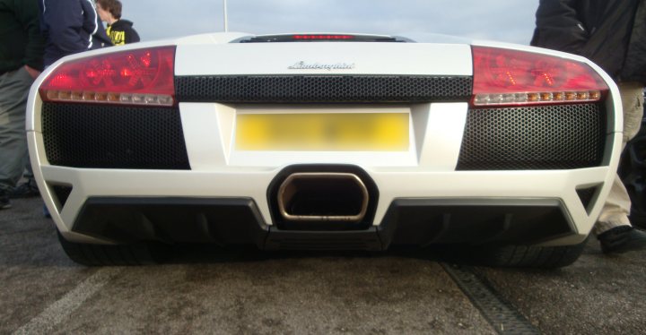 Oxford Service Pistonheads Sunday - The image features a white sports car with an aggressive rear spoiler. The car is sitting on a flat surface, and its distinctive tail lights are visible. There appears to be a person standing in the background, partially obscured by the car. The setting seems to be an outdoor area, possibly a parking lot, given the presence of what looks like a gray striped pole to the right. The lighting suggests it might be daytime.