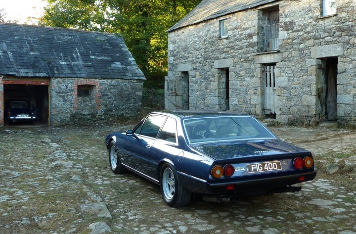 A mate's new toy - Page 1 - Ferrari V12 - PistonHeads - The image features an older, dark blue car parked in a gravel lot next to several stone buildings. The car appears to be a Datsun 240 Z model, which is a classic Japanese sports car. The buildings have a rustic appearance with stone walls and metal barred windows, suggesting they might be abandoned or repurposed structures. The scene exudes a sense of solitude and tranquility, with the car as the main point of interest. There is a hint of sunlight shining on the car, casting soft reflections on its shiny metal body.