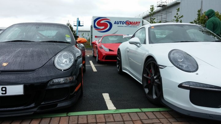 A car is parked on the side of the road - Pistonheads - In the image, a person can be seen in green attire to the right, standing near the Porsche car with a large blue and white text logo on the back of the vehicle. The Porsche and the asphalt-colored car, both sleek and shiny, are parked closely together, reflecting the commercial buildings in the background. The setting suggests a daytime street scene with a focus on the parked cars, one of which has its hood propped up by black, possibly auto-related, equipment on the floor behind the vehicle.