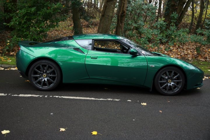 Might be an Evora owner soon - Page 3 - Evora - PistonHeads - The image showcases a sleek, green sports car parked on the side of a road on what appears to be an autumn day. The car is a two-door design, featuring a prominent rear spoiler, black wheels, and a low front bumper. The car's unique take on the traditional sports car design includes its streamlined shape and distinct shoulder drop characterizing the styling of the era. The backdrop is an avenue filled with trees, providing a natural splendor that contrasts with the car's modern, urban aesthetic. The reflections of light off the car's shiny surface give it a polished look, further adding to its overall appeal.